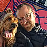 man, dog, person, selfie, portrait, beard, glasses, smiling, happy, tongue_out, long_hair, shaggy_dog, brown_dog, outdoor, sunlight, colorful_background, sign, street, jacket, passerby
