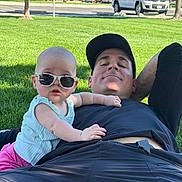 Lucy is registered to the contest to win money with this photo: baby, sunglasses, man, grass, park, outdoor, relaxing, casual, child, person, summer, greenery, shade, clothing, cap, resting, sunny, pink_pants, blue_top, smiling