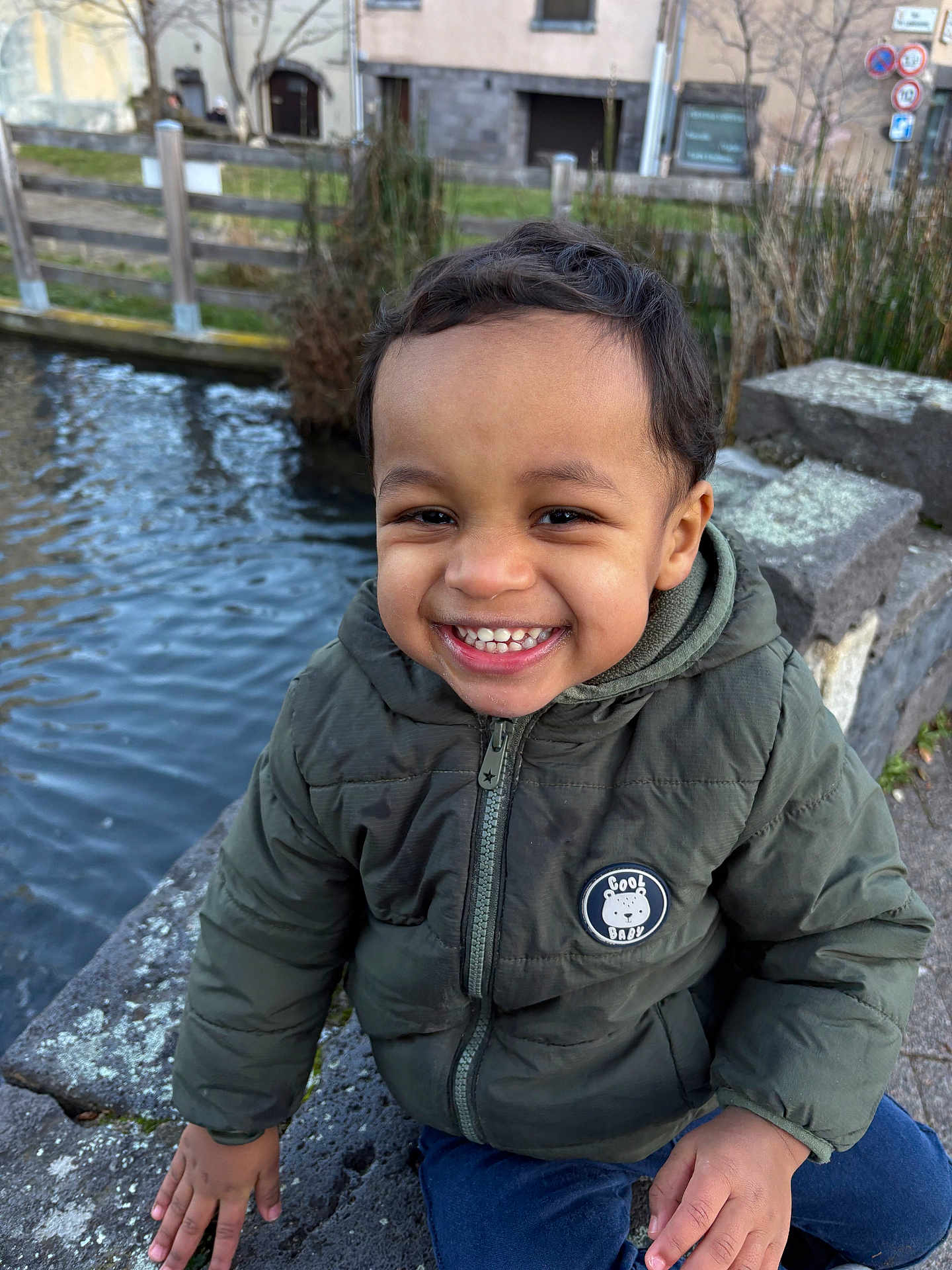 Karim participe au concours pour gagner de l'argent avec cette photo : toddler, child, smile, jacket, outdoor, water, canal, stone, hand, face, happy, winter_clothing, nature, park, fence, trees, urban, daylight, person, cute