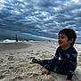 child, beach, sand, cloudy_sky, bird, ocean, water, smiling, sitting, outdoor, nature, waves, shoreline, dark_hair, long_sleeves, patterned_clothing, playful, relaxed, daytime, coastal