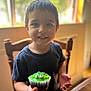 child, boy, cupcake, green_frosting, frosting_smudge, smile, face, short_hair, black_shirt, wooden_chair, table, indoor, natural_light, window, food, dessert, happy, portrait, cute, celebration