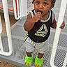 Romell is registered to the contest to win money with this photo: toddler, child, playground, play_equipment, slippers, green_slippers, clothing, shorts, tank_top, person, outdoor, sand, hand, face, hair, curious, grip, standing, toy, recreation