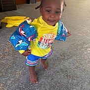 Romell joined the competition — help win amazing prizes! toddler, child, swimwear, floaties, shark_pattern, yellow_shirt, blue_shorts, barefoot, indoor, inflatable_toy, hair_braids, playful, smiling, person, floor, door, wall, colorful_clothing, cute, young_child