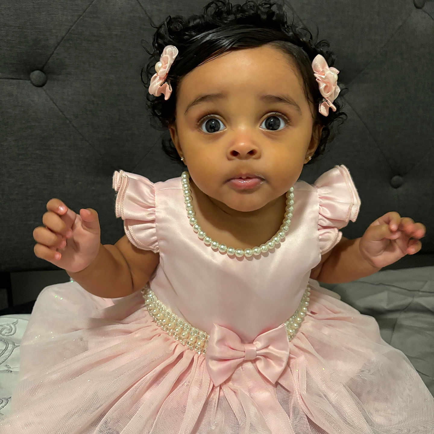 Alisha Roque joined the competition — help win amazing prizes! baby, child, portrait, pink_dress, pearl_necklace, flower_hair_clips, curly_hair, wide_eyes, sitting, smiling, indoors, tufted_backdrop, bed, formal_wear, jewelry, bow, ruffled_sleeves, tulle_skirt, hands, photography