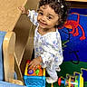 Alisha Roque joined the competition — help win amazing prizes! toddler, child, smiling, curly_hair, portrait, standing, indoors, playroom, toys, toy_train, colorful, play_mat, crib, furniture, table, chair, dress, floral_pattern, happy, 幼児