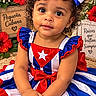 child, toddler, girl, portrait, smiling, curly_hair, bow, dress, patriotic, cuban, flag, red_white_blue, flowers, hibiscus, lantern, straw_hat, decorations, seated, indoors, festive