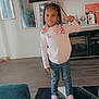 child, girl, braids, beads, peace_sign, smiling, casual_clothing, jeans, pink_shoes, living_room, television, rug, wood_floor, mirror, decor, photograph, furniture, home_interior, portrait, standing