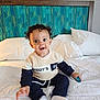 baby, child, curly_hair, sitting, bed, pillow, blanket, toy, clothing, sweater, pants, white_socks, indoor, headboard, blue, green, expression, surprise, delight, person