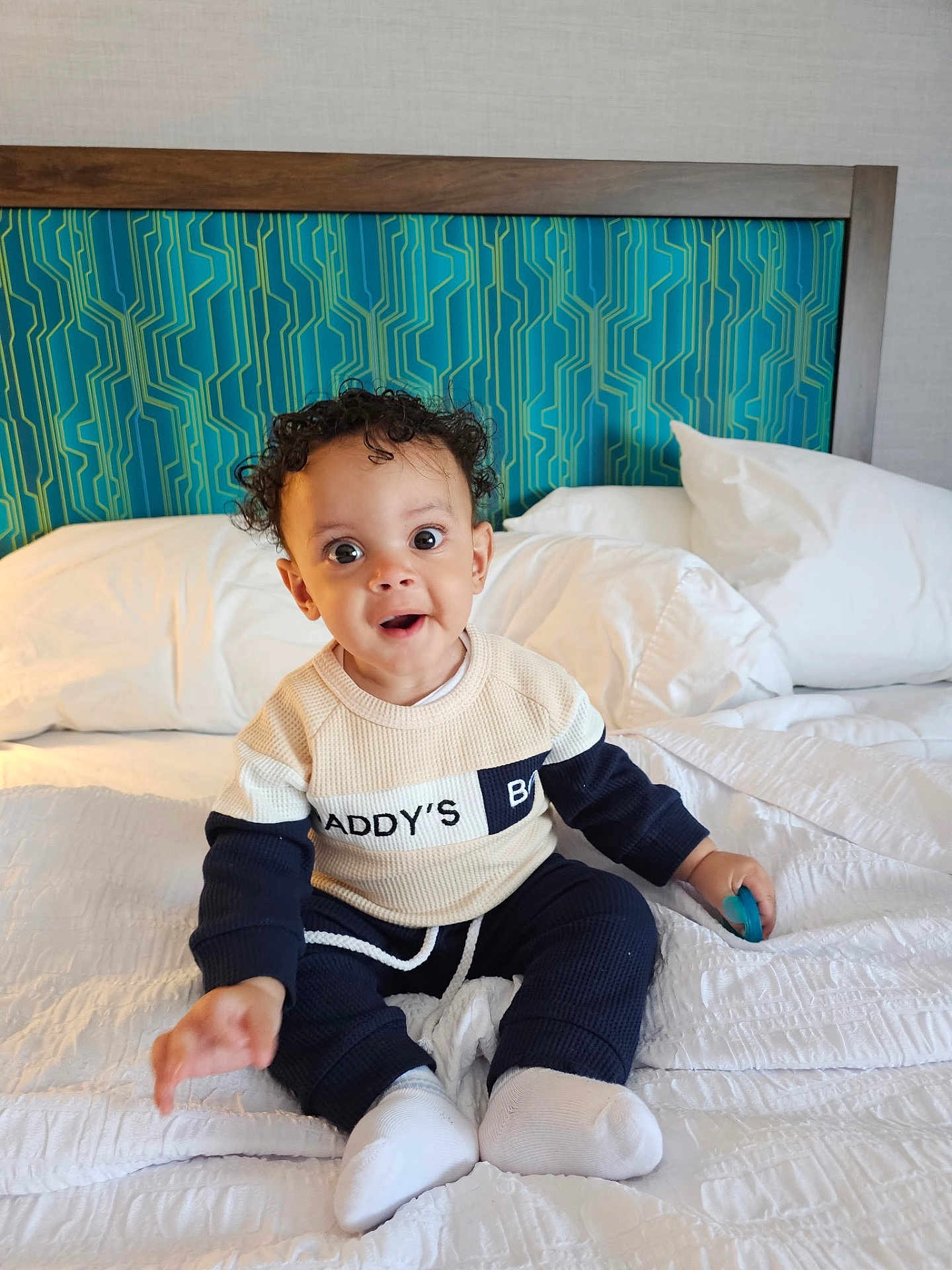 Arthur joined the competition — help win amazing prizes! baby, child, curly_hair, sitting, bed, pillow, blanket, toy, clothing, sweater, pants, white_socks, indoor, headboard, blue, green, expression, surprise, delight, person