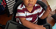 Jireh is registered to the contest to win money with this photo: toddler, child, high_chair, toy, plastic_egg, dinosaur_toys, instruction_sheet, striped_shirt, curly_hair, indoor, floor, cabinet, vaseline, vitamin_c, black_tray, table, hand, face, eyes, expression