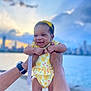 Brielle joined the competition — help win amazing prizes! baby, child, smiling, yellow_outfit, headband, adult_hands, waterfront, sunset, sky, clouds, city_skyline, outdoor, person, holding, cute, happy, infant, portrait, bright, daylight