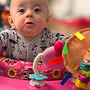 Silas joined the competition — help win amazing prizes! baby, infant, child, face, big_eyes, surprised_expression, onesie, printed_clothing, activity_center, toy, pink, playtime, indoors, portrait, shallow_depth_of_field, colorful, plastic_toy, seat, cute, closeup