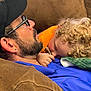 man, child, couch, curly_hair, glasses, cap, pacifier, blanket, orange_clothing, blue_clothing, brown_furniture, earring, facial_hair, resting, cuddling, indoor, close_up, comfort, nap, bonding