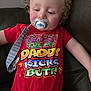 toddler, child, pacifier, red_shirt, curly_hair, sitting, couch, indoor, clothing, face, person, relaxed, cute, baby, infant, comfort, expression, colorful_text, strap, leisure