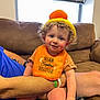 P.j joined the competition — help win amazing prizes! toddler, child, curly_hair, bib, orange_bib, knitted_hat, colorful_hat, indoor, couch, adult_arm, window, blinds, smiling, happy, person, seated, rosy_cheeks, casual_clothing, plaid_shorts, family