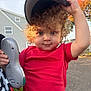 child, toddler, curly_hair, red_shirt, cap, toy, outdoor, daylight, house, tree, autumn, playful, person, face, hand, expression, street, casual_clothing, nature, holding