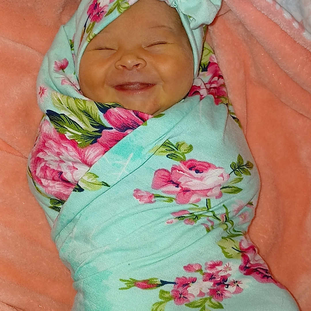 Liliana is registered to the contest to win money with this photo: baby, blanket, child, cozy, cute, fabric, face, floral_pattern, green, happy, headband, indoors, infant, newborn, person, pink, portrait, smile, soft_texture, wrapped