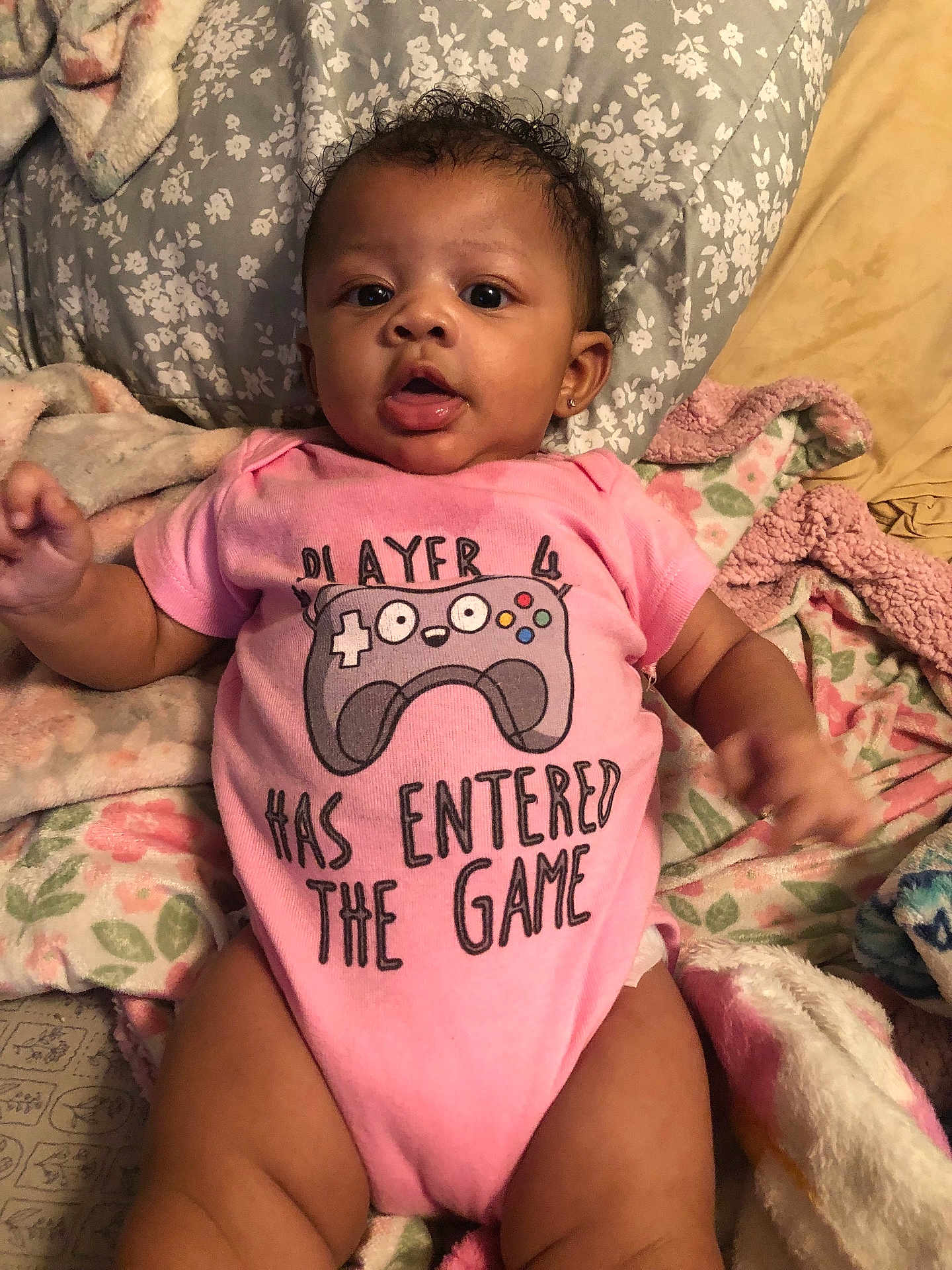 Hazellbelle joined the competition — help win amazing prizes! baby, infant, pink_onesie, gaming_controller, text_on_clothing, blanket, bed, floral_pattern, cute, child, portrait, face, expression, soft_texture, lying_down, indoors, comfort, cozy, small_child, casual