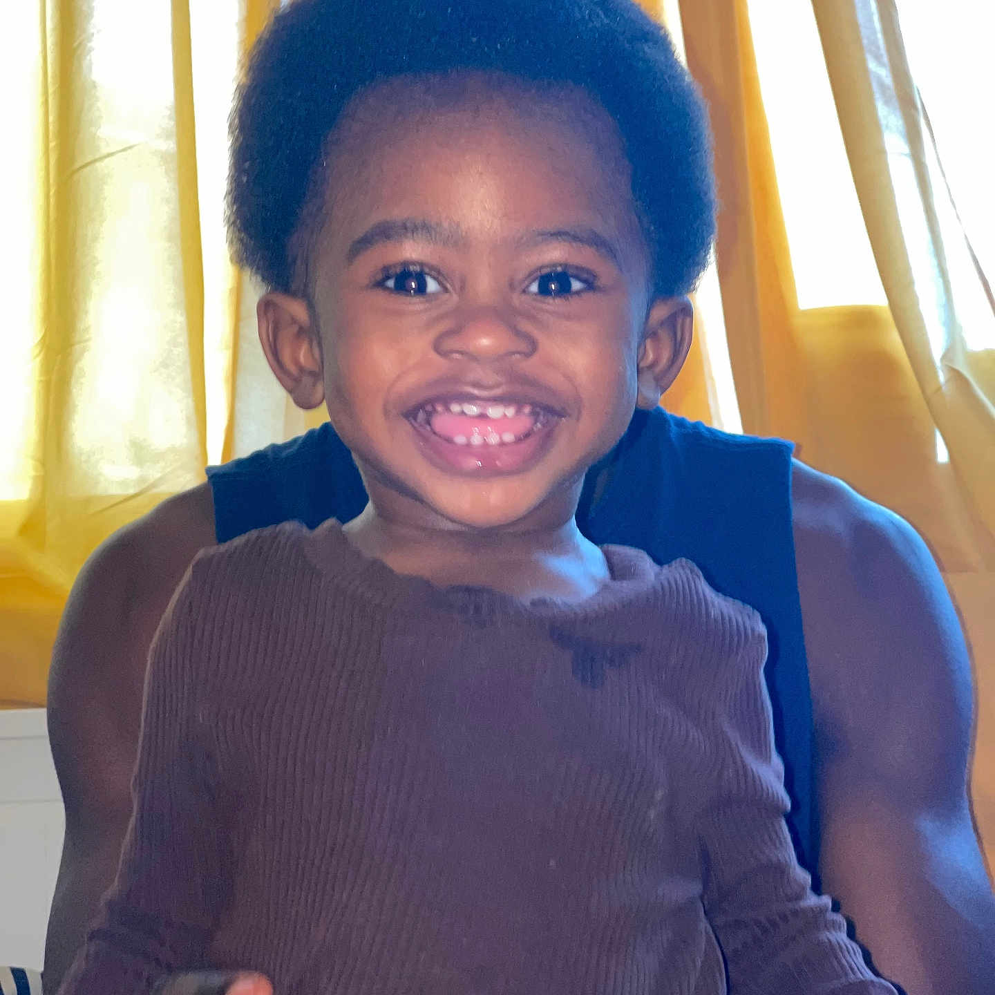 Moyi a rejoint le concours — aidez-le/la à gagner de superbes lots ! adult, afro_hair, background, brown_shirt, child, clothing, curtains, face, hands, happy, holding, indoor, light, long_sleeve, person, portrait, skin, smiling, toddler, window