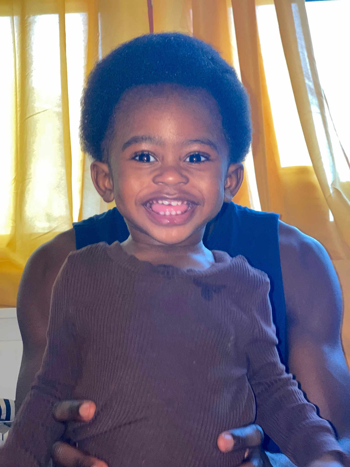 Moyi a rejoint le concours — aidez-le/la à gagner de superbes lots ! child, smiling, toddler, person, indoor, curtains, adult, holding, happy, face, clothing, long_sleeve, afro_hair, brown_shirt, hands, window, light, portrait, background, skin