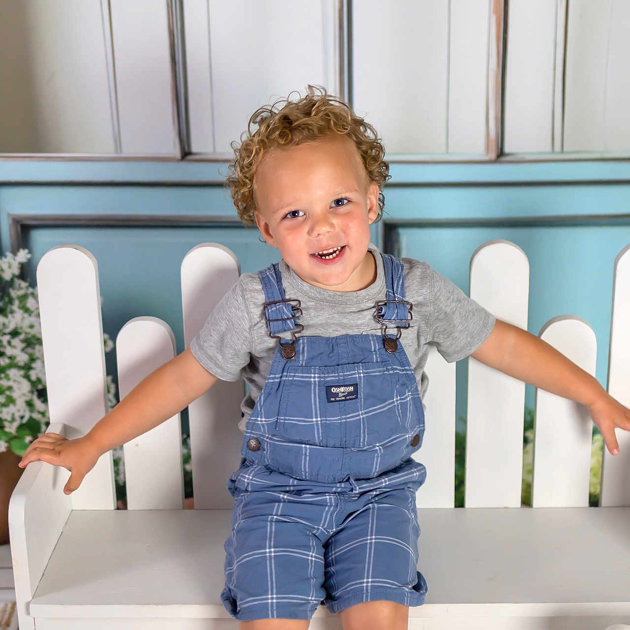 Jacoby joined the competition — help win amazing prizes! arms_outstretched, bench, blue_background, child, curly_hair, cute, flower_pot, flowers, grey_tshirt, happy, indoor, overalls, plaid, portrait, seated, smiling, toddler, white_bench, wooden_floor, young_child