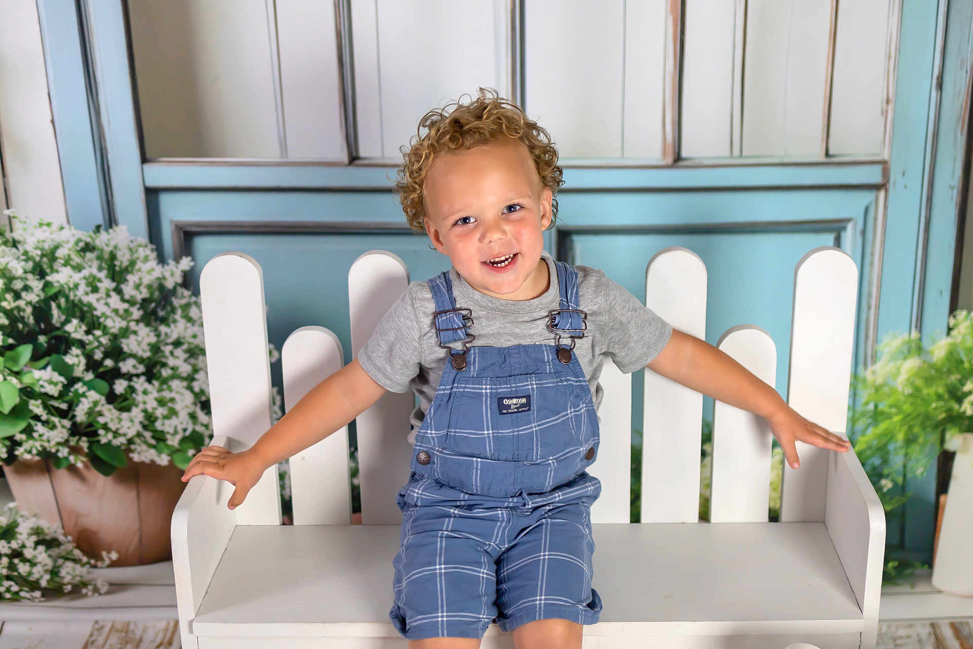 Jacoby joined the competition — help win amazing prizes! child, toddler, curly_hair, smiling, bench, overalls, grey_tshirt, white_bench, flower_pot, flowers, blue_background, wooden_floor, happy, indoor, portrait, cute, arms_outstretched, plaid, young_child, seated