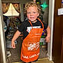 Jacoby is registered to the contest to win money with this photo: child, apron, home_depot, indoor, curtains, carpet, sneakers, black_tshirt, smiling, blond_hair, curly_hair, doorway, flooring, toy, holiday_decor, person, young_child, room, blurred_background, playful