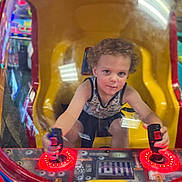 Jacoby joined the competition — help win amazing prizes! child, arcade_game, joystick, toddler, curly_hair, yellow_seat, red_lights, gaming, indoor, glass_reflection, bright_colors, playful, fun, young_child, concentration, entertainment, technology, amusement, machine, seat