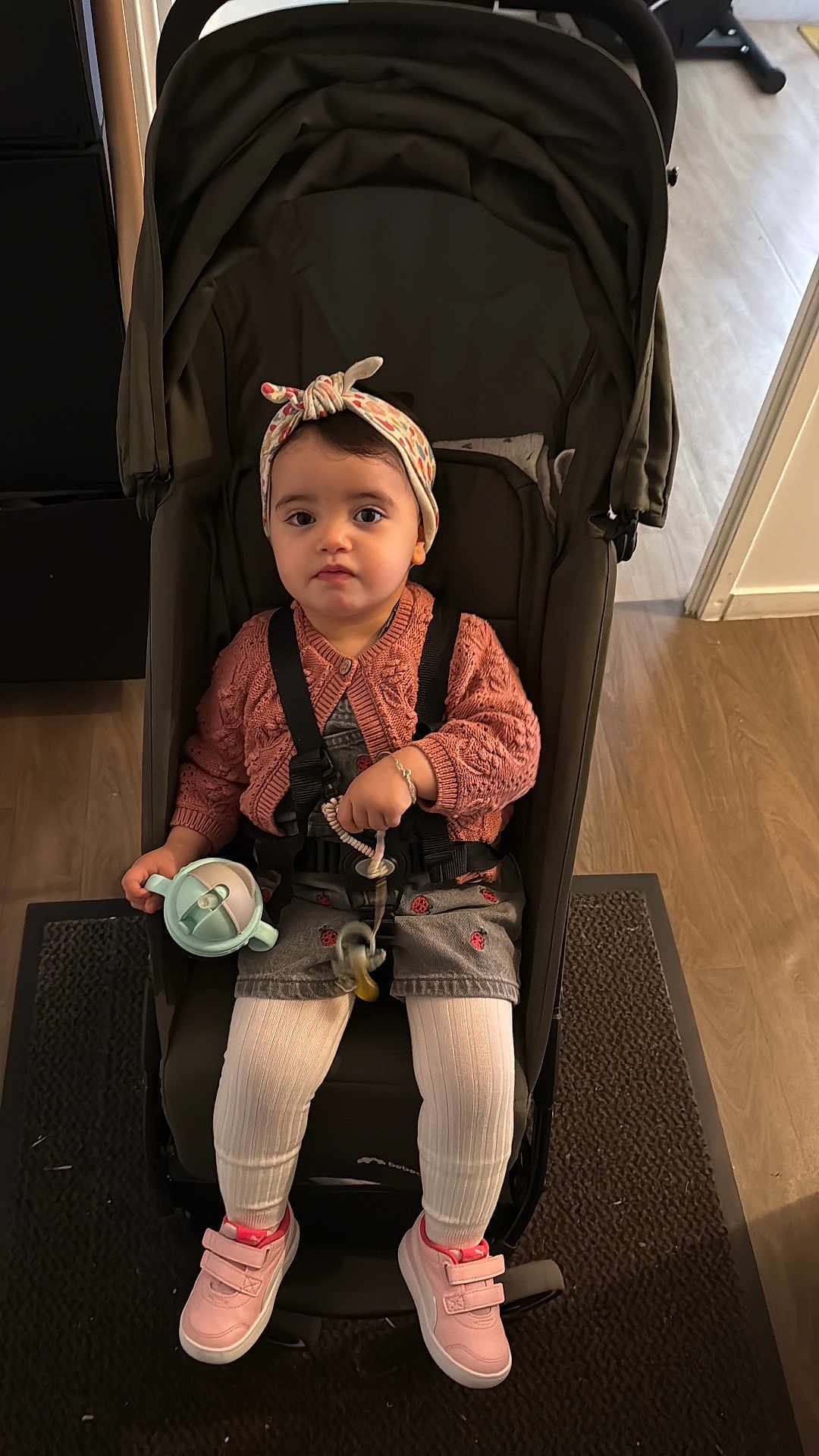 Mariame a rejoint le concours — aidez-le/la à gagner de superbes lots ! toddler, child, stroller, pink_cardigan, headband, pacifier, sippy_cup, white_tights, pink_shoes, indoor, carpet, wooden_floor, bracelet, curly_hair, seated, baby, young_child, person, cute, casual_clothing