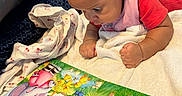 Kailani is registered to the contest to win money with this photo: baby, child, reading, book, picture_book, blanket, pillow, indoor, curious, toddler, colorful, illustration, soft_fabric, cozy, lying_down, infant, clothing, focus, early_learning, home