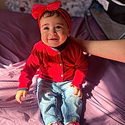 Zainab Samhan joined the competition — help win amazing prizes! baby, child, smiling, red_sweater, headband, jeans, shoes, indoor, bed, hand, person, happy, cute, portrait, sitting, sunlight, clothing, footwear, infant, casual