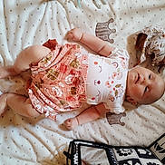Lena participe au concours pour gagner de l'argent avec cette photo : baby, infant, blanket, plush_toy, clothing, patterned, cute, lying_down, smiling_face, soft_texture, child, person, indoor, skirt, white_shirt, toy, bag, decor, relaxed, happy