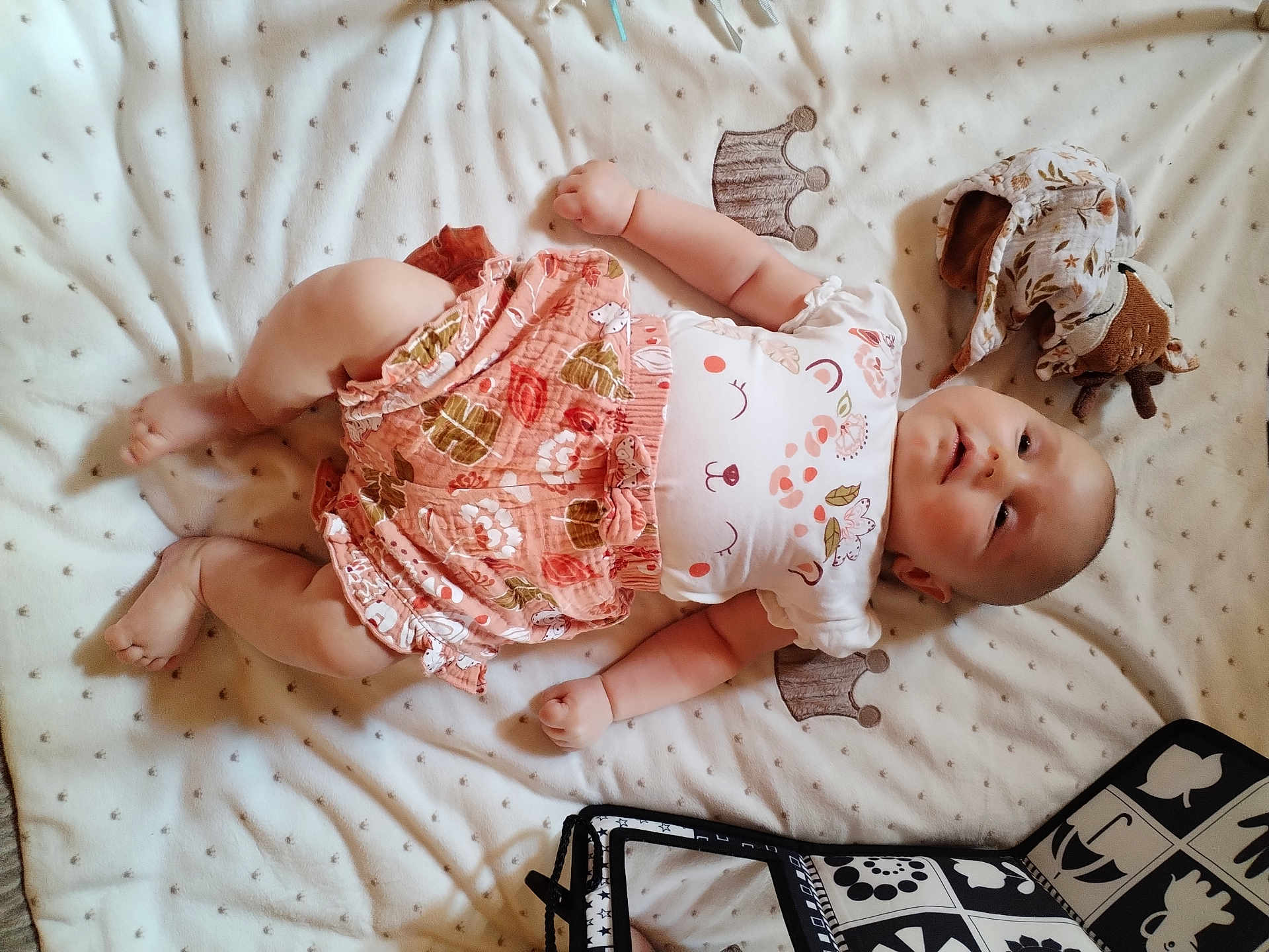 Lena participe au concours pour gagner de l'argent avec cette photo : baby, infant, blanket, plush_toy, clothing, patterned, cute, lying_down, smiling_face, soft_texture, child, person, indoor, skirt, white_shirt, toy, bag, decor, relaxed, happy