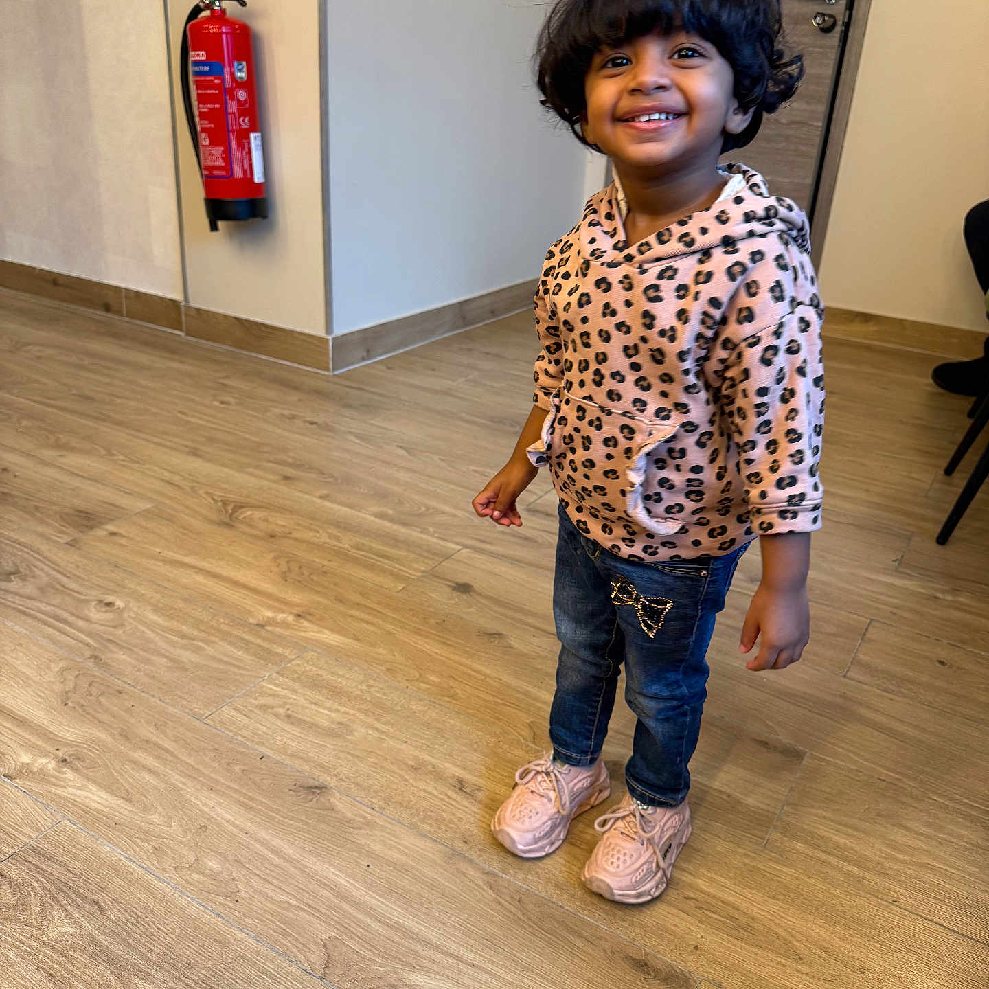 Faiha participe au concours pour gagner de l'argent avec cette photo : casual_clothing, child, curly_hair, cute, door, fire_extinguisher, floor, happy, hoodie, indoor, interior, jeans, leopard_print, person, portrait, smiling, sneakers, standing, wooden_floor, young_child
