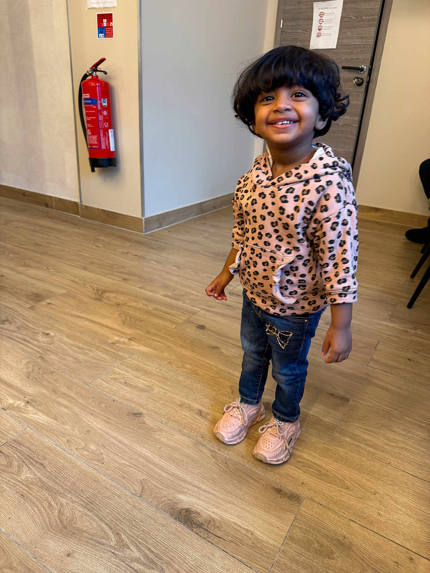 Faiha participe au concours pour gagner de l'argent avec cette photo : child, smiling, indoor, wooden_floor, hoodie, leopard_print, jeans, sneakers, fire_extinguisher, door, person, happy, standing, curly_hair, casual_clothing, floor, interior, young_child, cute, portrait