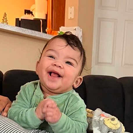 Liam is registered to the contest to win money with this photo: baby, child, smiling, laughing, green_clothing, indoor, living_room, couch, plush_toy, happy, cute, person, infant, head, face, home, cozy, striped, furniture, light