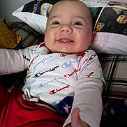 Stephen is registered to the contest to win money with this photo: baby, smiling, tongue_out, champion_clothing, red_pants, white_shirt, pillow, bed, blanket, indoor, happy, child, person, infant, cute, reaching_hand, face, head, cozy, portrait