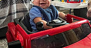 Stephen is registered to the contest to win money with this photo: baby, blanket, blue_shirt, car_seat, child, cute, dog, furniture, happy, helmet, indoor, lamp, living_room, photograph, playtime, red, smile, steering_wheel, toy, toy_car