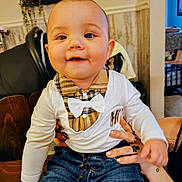 Stephen is registered to the contest to win money with this photo: adult_hand, baby, bow_tie, cheeks, child, eyes, face, hands, highchair, home_interior, indoor, infant, jeans, plaid, plant, portrait, shirt, smiling, wall_decor, wooden_table