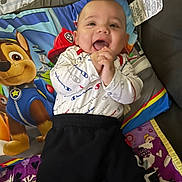 Stephen is registered to the contest to win money with this photo: baby, child, smiling, clapping, paw_patrol, pillow, blanket, clothing, white_shirt, black_pants, happy, indoors, cute, infant, person, colorful, playful, soft, cozy, portrait