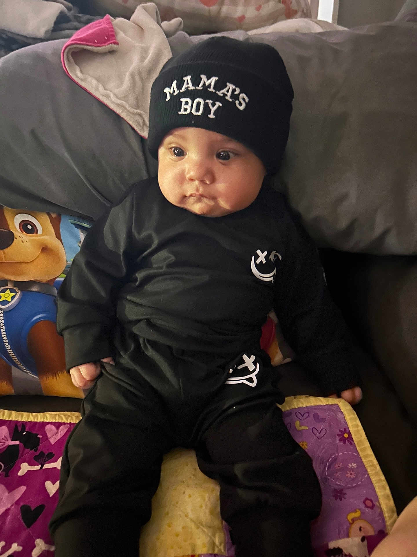 Stephen joined the competition — help win amazing prizes! baby, child, black_clothing, beanie, hat, sitting, quilt, pillow, paw_patrol, indoors, cute, chubby_cheeks, face, person, clothing, blanket, expression, portrait, young_child, casual