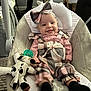 baby, child, infant, smiling, tongue_out, headband, bow, plaid, overalls, stuffed_toy, cow_toy, pacifier, baby_seat, cushion, indoor, carpet, furniture, person, happy, cute