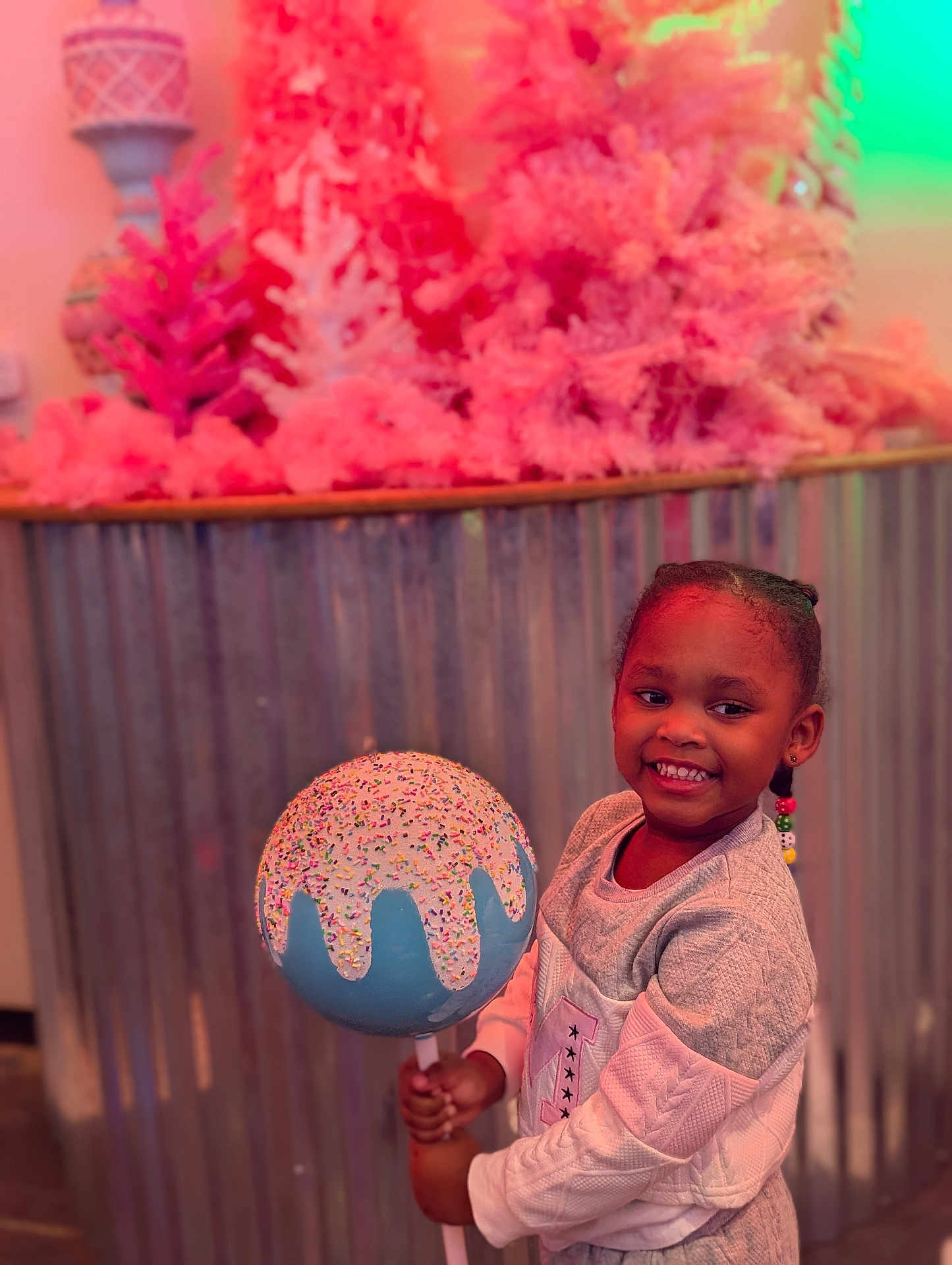 Coirey joined the competition — help win amazing prizes! child, girl, smile, lollipop, sprinkles, candy, braids, beads, sweater, portrait, indoor, decor, pink_trees, corrugated_metal, background, holding, happy, colorful, playful, props