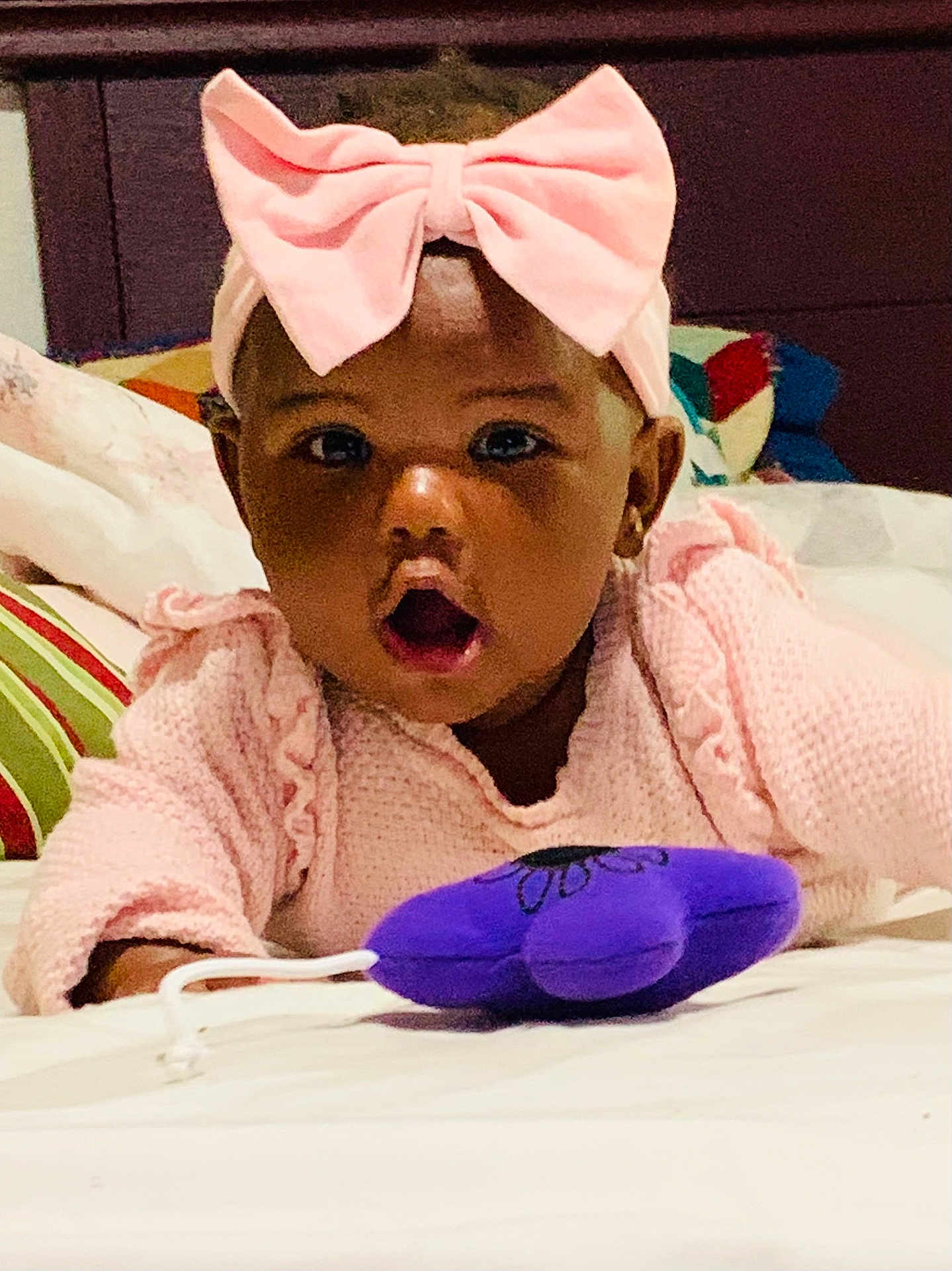 Alexandria joined the competition — help win amazing prizes! baby, child, pink_bow, headband, pink_sweater, purple_toy, plush_toy, bed, pillow, curious, surprised, indoor, cozy, face, person, infant, cute, expression, childhood, soft_toy