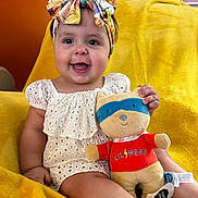 Adeline is registered to the contest to win money with this photo: baby, child, headband, toy, plush_toy, smiling, sitting, yellow_blanket, orange_wall, cute, infant, happy, indoors, face, clothing, person, soft_toy, colorful, portrait, young_child