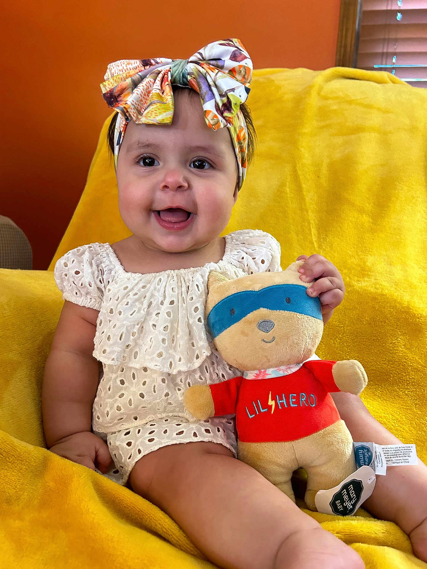 Adeline is registered to the contest to win money with this photo: baby, child, headband, toy, plush_toy, smiling, sitting, yellow_blanket, orange_wall, cute, infant, happy, indoors, face, clothing, person, soft_toy, colorful, portrait, young_child