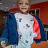 child, toddler, smiling, indoor, jacket, mickey_mouse, clothing, necklace, cross, face, person, cute, happy, table, chair, ceiling, light, window, orange_wall, pants