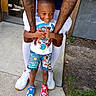 child, boy, smiling, tattoos, colorful_clothing, paw_patrol, shorts, sneakers, adult, arms, outdoor, concrete, door, plants, happy, playful, holding, feet, casual, summer