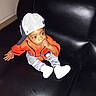 Cassius joined the competition — help win amazing prizes! toddler, child, baseball_cap, orange_jersey, striped_pants, white_shoes, couch, leather, indoor, sitting, thumb_in_mouth, wide_eyes, curious, casual_clothing, young_child, portrait, fashion, hat, sportswear, black_background