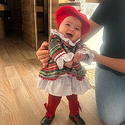 Lïya participe au concours pour gagner de l'argent avec cette photo : baby, child, smiling, red_beret, plaid_dress, indoor, wooden_floor, adult_hand, supporting, happy, footwear, red_tights, person, flooring, home_decor, vase, plant, kneeling, cute, portrait